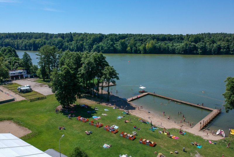 Uferwiese mit Liegestühlen, Sandstrand und eingezäuntem Badepier; Gäste sonnen sich und schwimmen auf einem ruhigen, waldumsäumten See