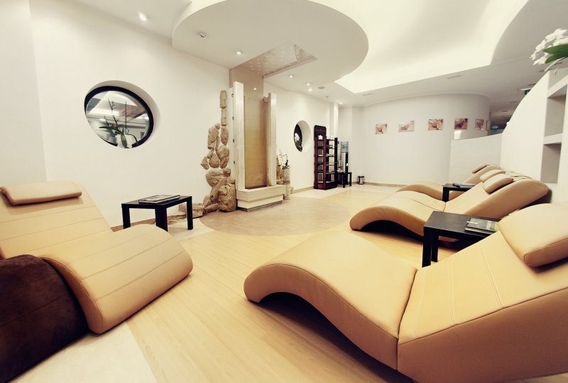 Ruheraum mit geschwungenen beigen Liegen, Innenwasserfall, sanfter Beleuchtung und Beistelltischen.