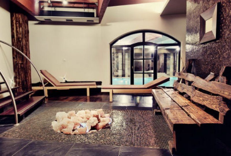 Spa-Lounge mit Holzbank und Liegen, Salzstein-Dekor auf Mosaikboden und bogenförmigen Glastüren zum Hallenbad.