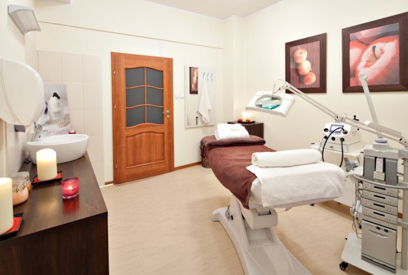 Ruhiger Behandlungsraum mit verstellbarer Behandlungsliege, Lupenlampe, Kosmetikgeräten, Duftkerzen und Waschbecken.