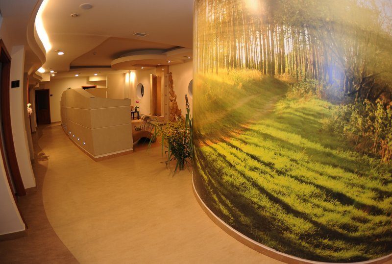 Geschwungener Hotelflur mit moderner Rezeption, warmen Deckenleuchten und großem, sonnenhellen Waldwandbild in Spa‑Atmosphäre