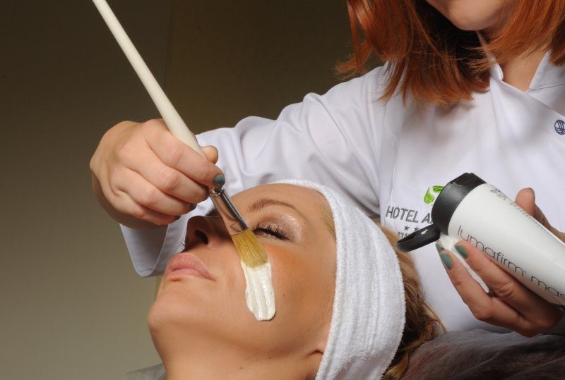 Gast erhält cremige Gesichtsmaske, mit Pinsel aufgetragen von Therapeutin in weißer Dienstkleidung im Hotelspa.