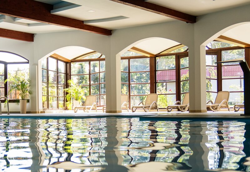 Überdachtes Hotel-Schwimmbad mit bogenförmigen Panoramafenstern, sonnigen Liegen, grünen Topfpflanzen und sichtbaren Holzdeckenbalken