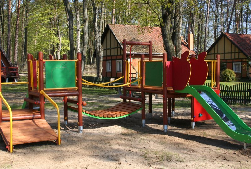 Kinderspielplatz mit grüner Rutsche, Holzsteg und Spieltafeln neben Fachwerk-Ferienhäusern im schattigen Waldgrundstück