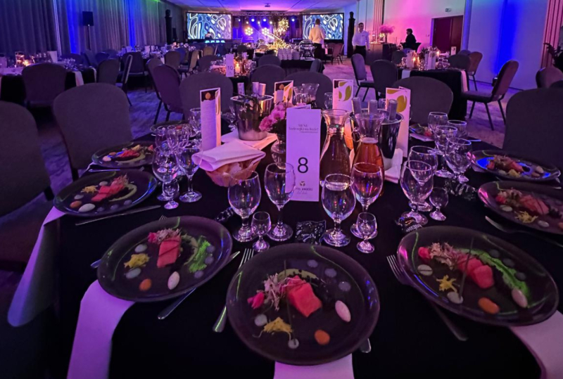 Banketttisch gedeckt für Gala-Dinner mit angerichteten Vorspeisen, mehreren Weingläsern, Blumen und violetter Bühnenbeleuchtung.