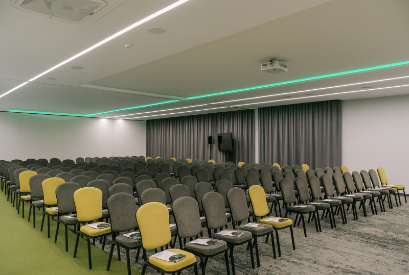 Moderner Hotelkonferenzraum mit Reihen grauer und gelber Stühle, Deckenprojektor, grünem LED-Licht und Vorhangleinwand.