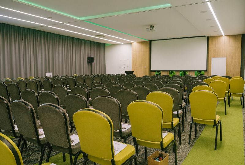 Hotel-Konferenzraum mit Reihen grauer und limonengrüner Stühle, Projektionsleinwand, Rednerpult, LED-Deckenbeleuchtung und grünem Teppich