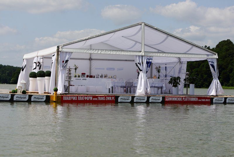 Weißes, offenes Veranstaltungszelt auf einer schwimmenden Plattform über einem ruhigen See, mit Buffet, eingedeckten Tischen, weißen Stühlen und Topfpflanzen