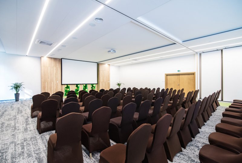 Moderner Konferenzraum im Hotel mit Reihen brauner Stühle vor großer Projektionsleinwand, Teppichboden und gedämpftem Licht.
