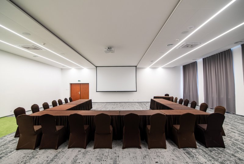 Geräumiger Hotelkonferenzraum mit U-förmigen Tischen in braunen Hussen, Leinwand, Deckenbeleuchtung und hohen Vorhängen