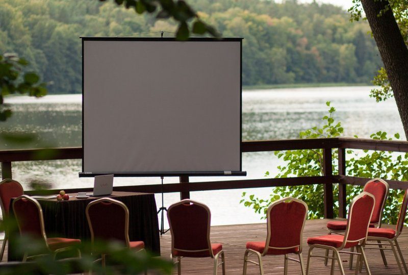 Holzterrasse am See für Tagung mit Projektionsleinwand, roten Stühlen und Tisch mit Laptop und Äpfeln, ruhige Wald- und Wasseransicht