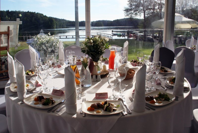 Festlich gedeckter Tisch am See mit weißen Tischdecken, Blumenarrangement, aufgestellten Servietten, angerichteten Vorspeisen und Gläsern, Blick auf einen Holzsteg.