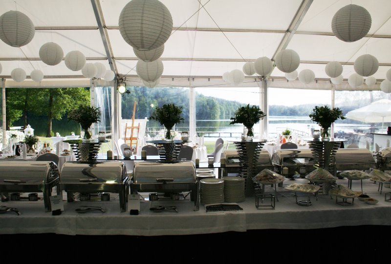 Buffet im weißen Festzelt am See mit Warmhaltern, Papierlaternen, Blumenarrangements und gedeckten Tischen für ein festliches Essen