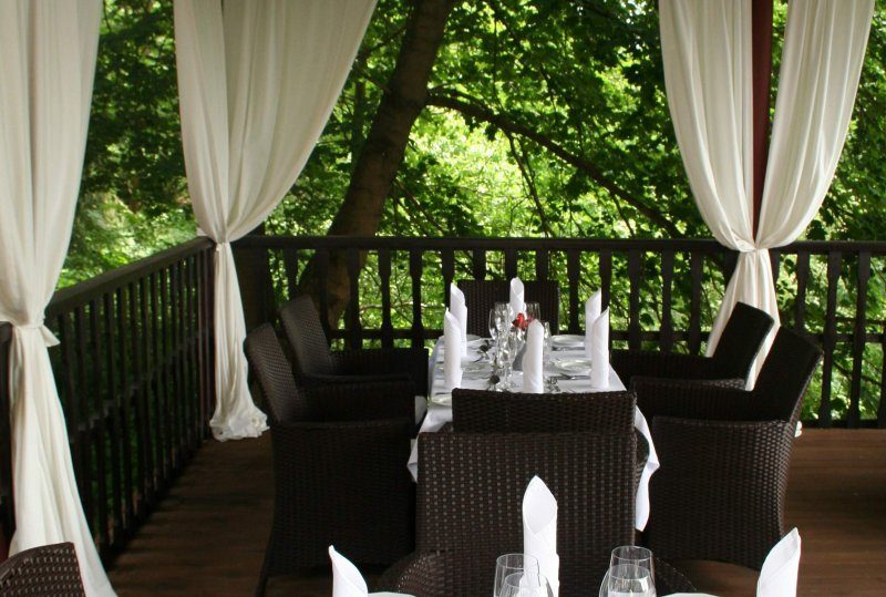 Überdachte Terrasse zum Essen mit Rattanstühlen, weiß gedecktem Tisch, Gläsern und Vorhängen, mit Blick auf grünen Wald.