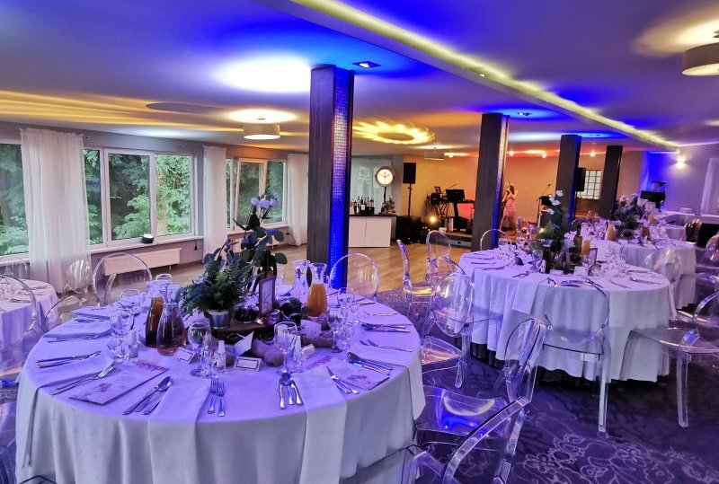 Bankettraum mit runden Tischen in weißen Tischdecken, Blumenarrangements, transparenten Stühlen, farbiger Beleuchtung, Bar und Bühne