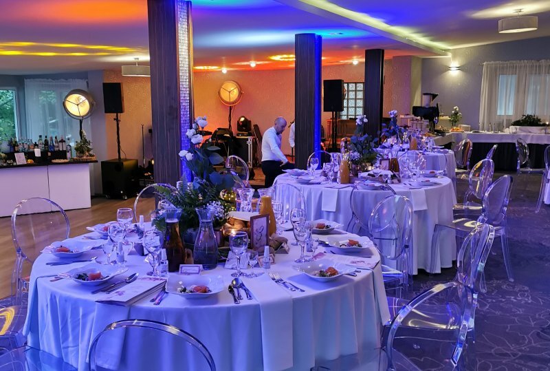 Bankettraum mit runden Tischen, weißen Tischdecken, Blumenarrangements, transparenten Stühlen, farbigem Stimmungslicht und Personal.