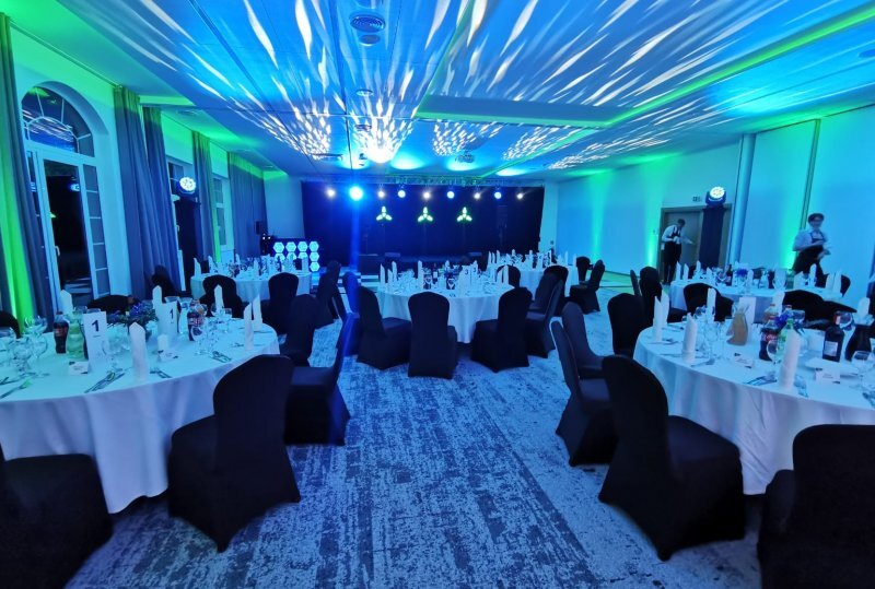 Hotelballsaal fürs Bankett mit runden Tischen, Tischdecken, schwarzen Stuhlhussen und Gedeck; Personal bereitet Bühne mit blau-grünem Licht vor.