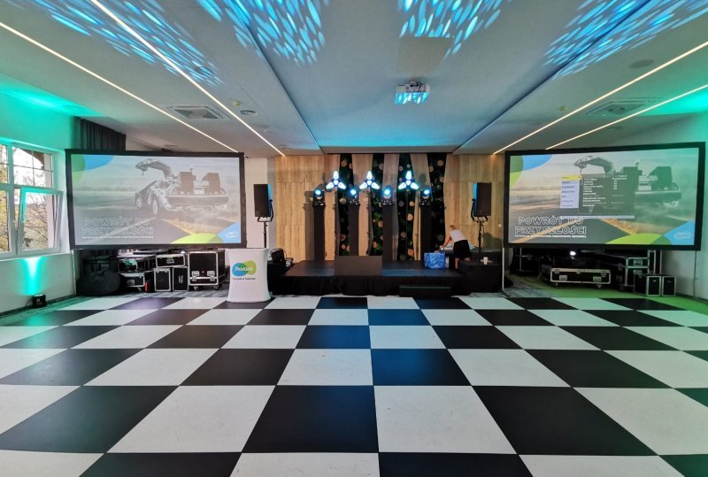 Veranstaltungsraum mit schwarz-weiß kariertem Tanzboden, erhöhter Bühne mit Beleuchtung, zwei Projektionsleinwänden und türkisfarbener Akzentbeleuchtung; Personal bereitet Bühne vor.