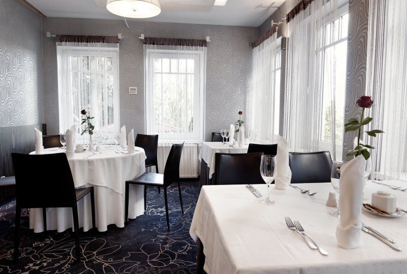 Kleines Hotelrestaurant mit weißen Tischdecken, gefalteten Servietten, Weingläsern und einzelnen roten Rosen an großen Fenstern mit Vorhängen.