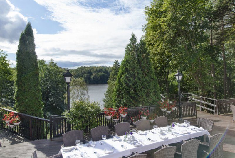 Holzterrasse am See mit langem weiß gedecktem Tisch, Korbsesseln, Topfblumen, Laternen und Bäumen, ruhiger Seeblick