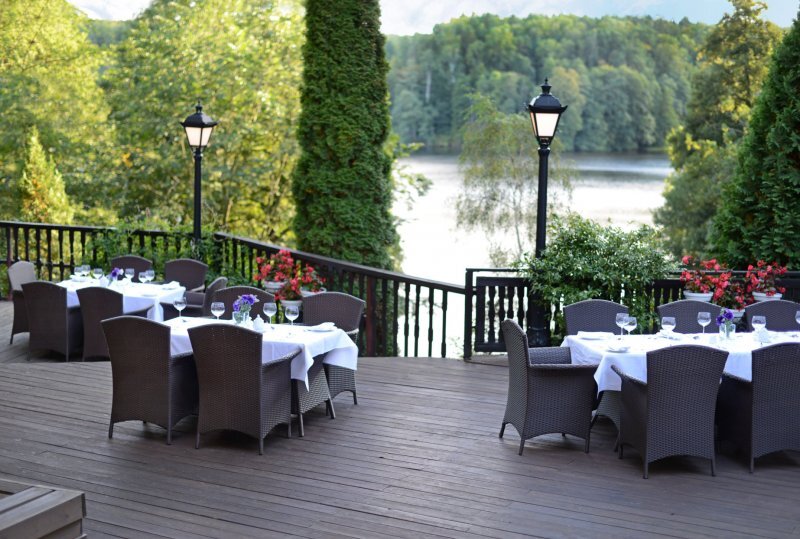 Restaurantterrasse am See mit Holzdeck, Rattanstühlen, weißen Tischdecken, Laternen und grünem Waldblick.
