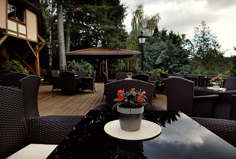 Hotelterrasse mit Rattansesseln, glänzendem Tisch mit Topf roter Blumen, Holzdeck, Pavillon und Bäumen.