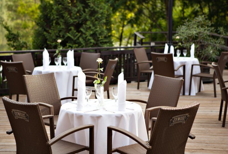 Restaurantterrasse auf Holzdeck mit runden Tischen in weißen Tischdecken, gefalteten Servietten, Weingläsern und Rattanstühlen vor grünem Laub