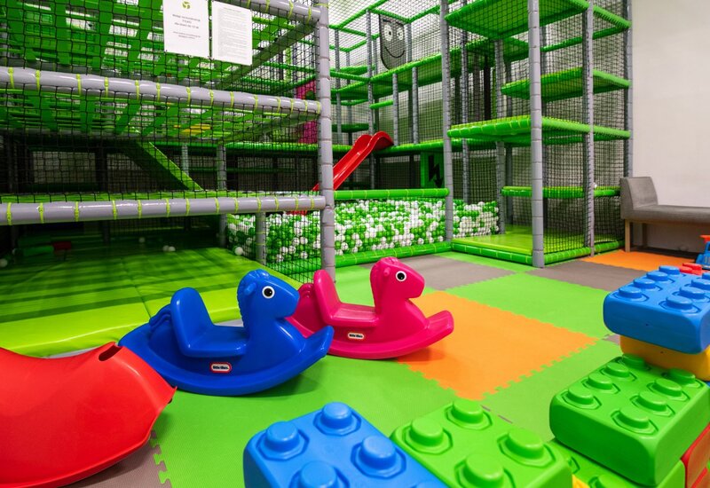 Überdachter Kinder-Spielbereich mit buntem Schaumstoffboden, zwei Plastikwippen, großen Bausteinen, mehrstöckigem Klettergerüst und Bällebad