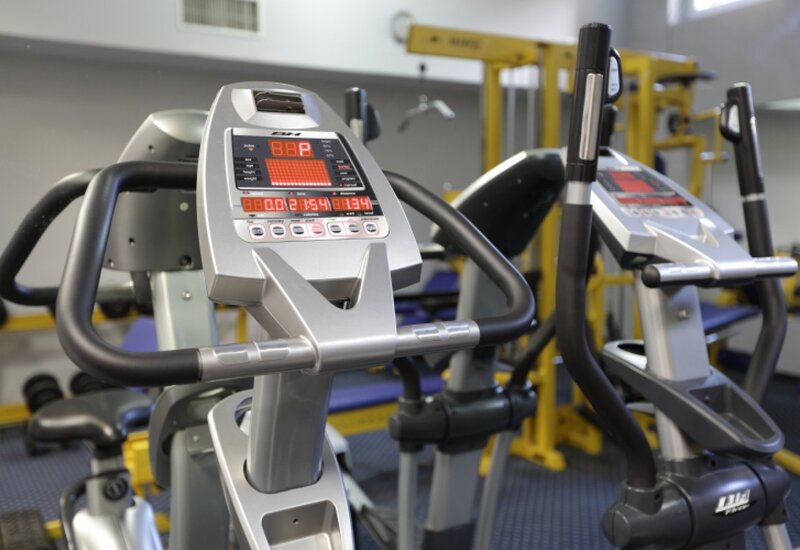 Hotel-Fitnessraum mit modernen Crosstrainern, digitalen Konsolen und gelbem Hantelgestell.