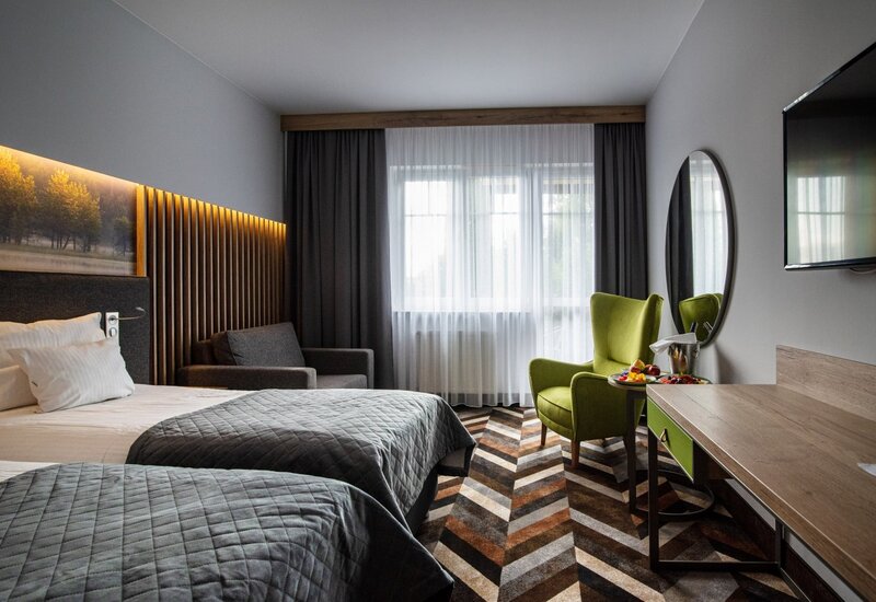 Modernes Hotelzimmer mit zwei Betten, hinterleuchtetem Holz-Kopfteil, grünem Sessel, Schreibtisch und TV, Fenster mit Gardinen, Obst und Champagnerkühler