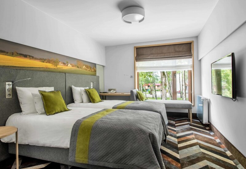 Modernes Zweibettzimmer mit grauen Bettdecken und grünen Akzenten, Fensterbank mit Blick auf Bäume und Wand-TV