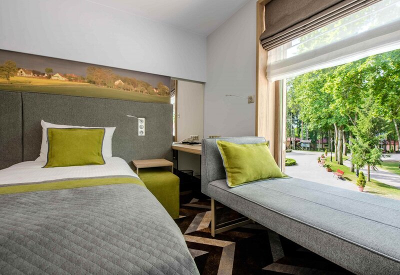 Einzelzimmer mit grauem Bett und Liege, grünen Kissen, Schreibtisch und großem Fenster mit Blick auf einen baumbestandenen Park und Bänke.