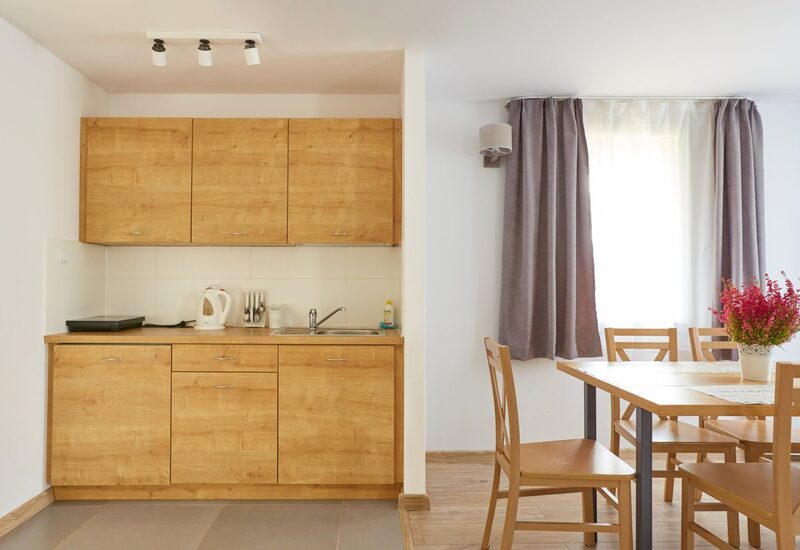 Hotel-Küchenzeile mit warmen Holzschränken, Spüle, Wasserkocher und Kochfeld; Esstisch mit Stühlen und Blumenarrangement am hellen Fenster.