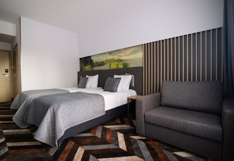 Zwei Einzelbetten mit grauen Tagesdecken, Landschaftsbild überm Kopfteil, Nachttisch und dunkelgraues Sofa in modernem Hotelzimmer.