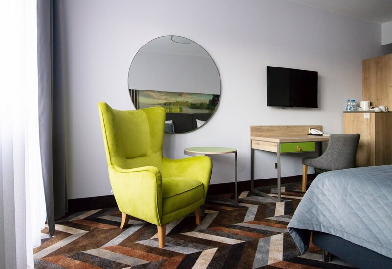 Helles modernes Hotelzimmer mit limonengrünem Sessel, rundem Spiegel, Schreibtisch mit TV, Wasserkocher und gestepptem Bett