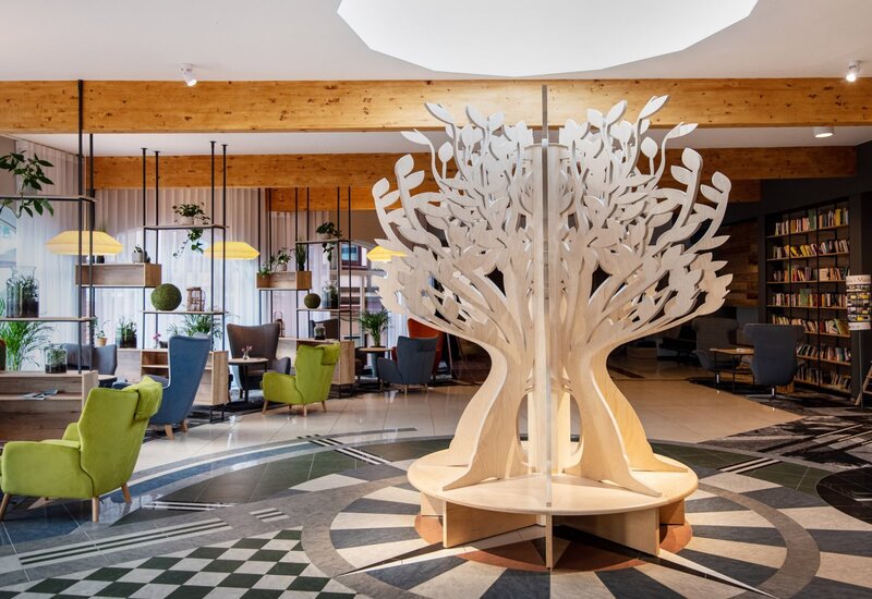 Skulpturales Holz-„Baum“-Sitzmöbel in der Hotellobby mit grünen und blauen Sesseln, Bücherregalen, Pflanzen und warmen Holzbalken
