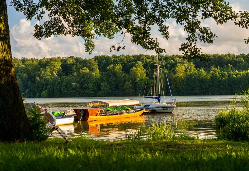 Holzboot mit Sonnendach und Segelboot an ruhigem See im goldenen Abendlicht, bewaldetes gegenüberliegendes Ufer und schattiges Grasufer