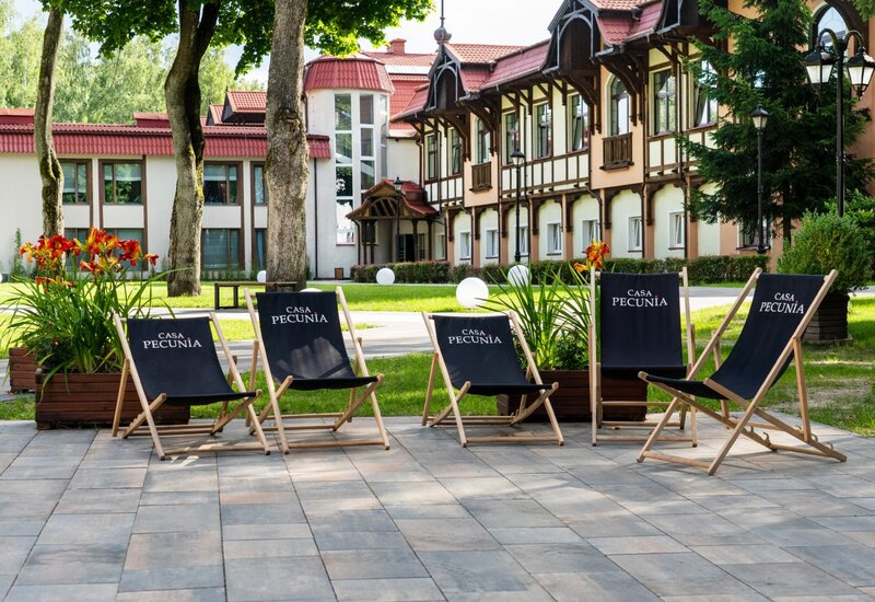 Casa Pecunia Liegestühle auf gepflasterter Terrasse, neben Rasen, Bäumen und blühenden Kübeln vor einem Hotel mit Holzfassade.