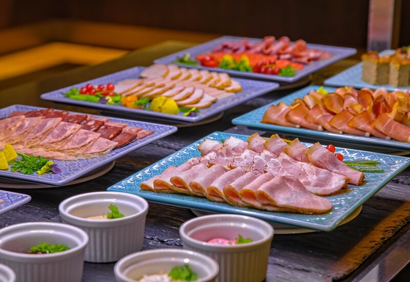 Frühstücksbuffet mit verschiedenen aufgeschnittenen Wurst- und Schinkensorten auf blauen Platten, garniert mit Kräutern, Cocktailtomaten und Soßenschälchen.