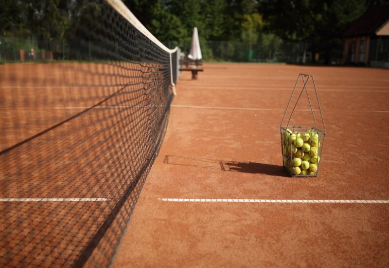 Clay tennis court with net in foreground, wire basket of yellow balls by the baseline, bench and umbrella in the background