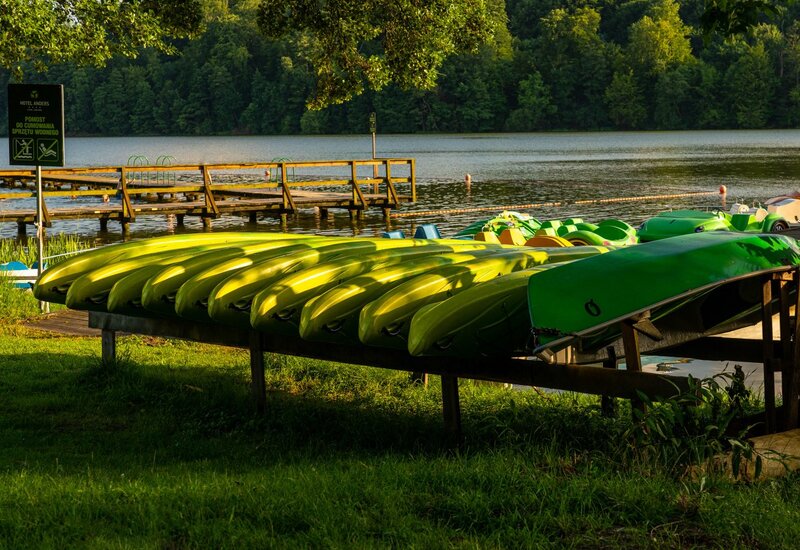 Helle limonengrüne Kajaks auf Gestell neben hölzernem Hotelsteg, ruhiger See und Wald im warmen Abendlicht