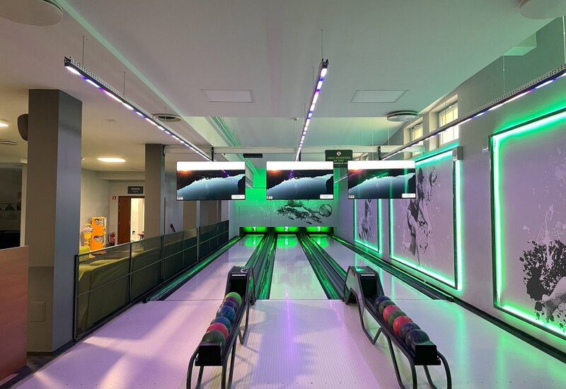 Moderne Bowlingbahn mit drei Bahnen, grünem Neonlicht, Anzeigemonitoren über den Bahnen und Kugelständern