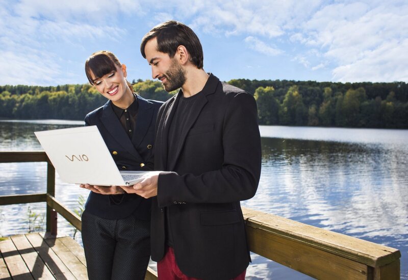 Gut gekleidetes Paar lächelt über einen Laptop auf einer hölzernen Hotelterrasse am See, mit Blick auf das bewaldete Ufer und einen Außenarbeitsplatz.