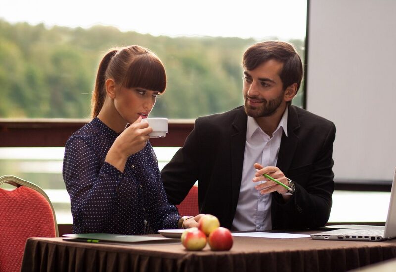 Zwei Kollegen an einem Hotel-Tagungstisch am Fenster; Frau trinkt Kaffee, Mann erklärt mit Stift, Laptop und Äpfel auf dem Tisch