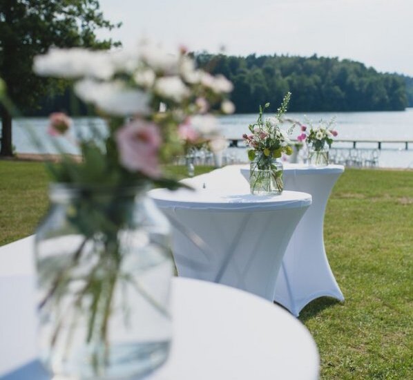 Stehtische am See mit weißen Hussen und Einmachglas-Blumenarrangements auf der Wiese, im Hintergrund Steg und bewaldetes Ufer