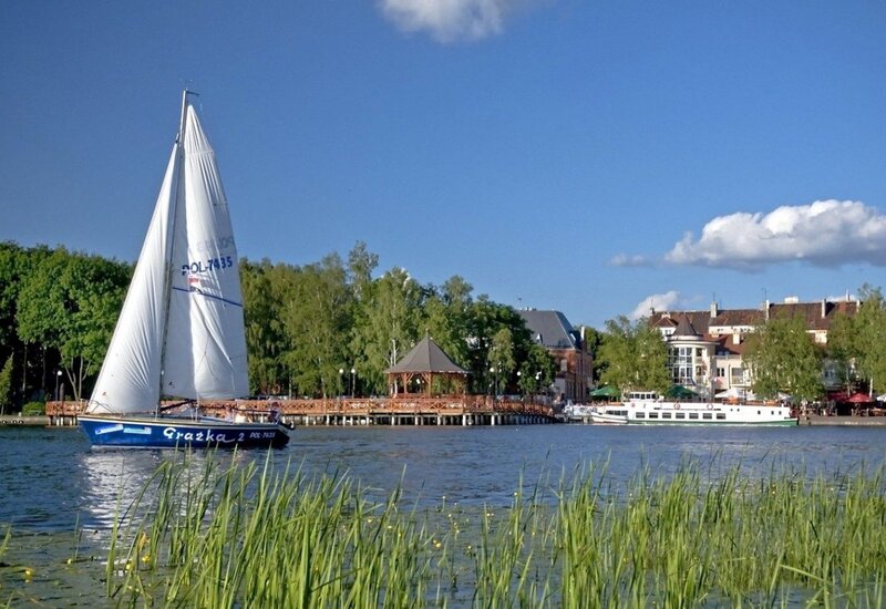 Segelboot auf dem See vor einem hölzernen Pavillon am Steg, Uferrestaurants und Hotelterrassen unter blauem Himmel.