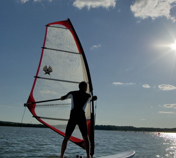 Gast windsurft auf ruhigem See mit rot akzentuiertem Segel, tief stehender Sonne und vereinzelten Wolken.
