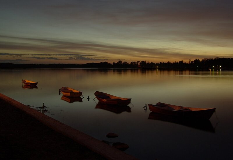 Ruderboote am ruhigen See bei Dämmerung, warme Sonnenuntergangsreflexe und bewaldetes Ufer.