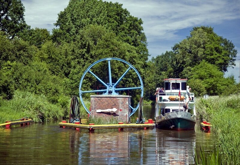Historisches blaues Wasserrad auf Ziegelplattform neben einem Ausflugsschiff im baumgesäumten Kanal, ruhige Spiegelung