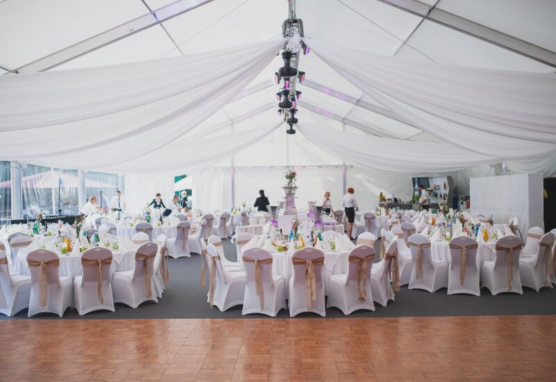 Elegantes weißes Festzelt mit drapiertem Dach, eingedeckte Rundtische für Hochzeit, Stühle mit beigen Schleifen und Blumenarrangements